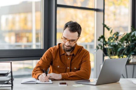 Empresário de barva usando óculos e camisa moderna escrevendo em caderno perto de notebook